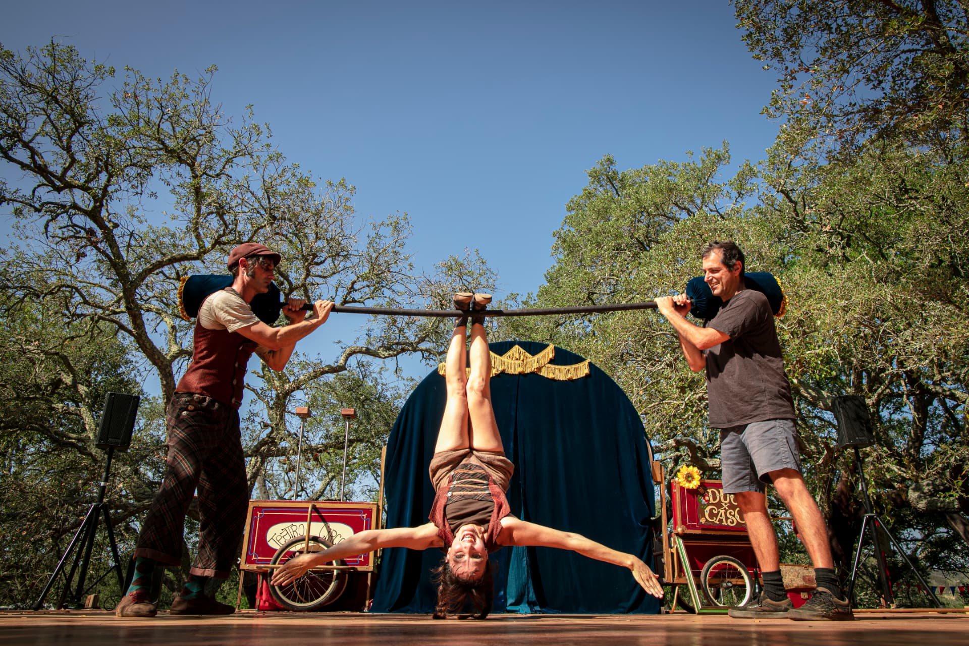 Acrobata suspensa numa barra horizontal entre dois artistas no palco ao ar livre