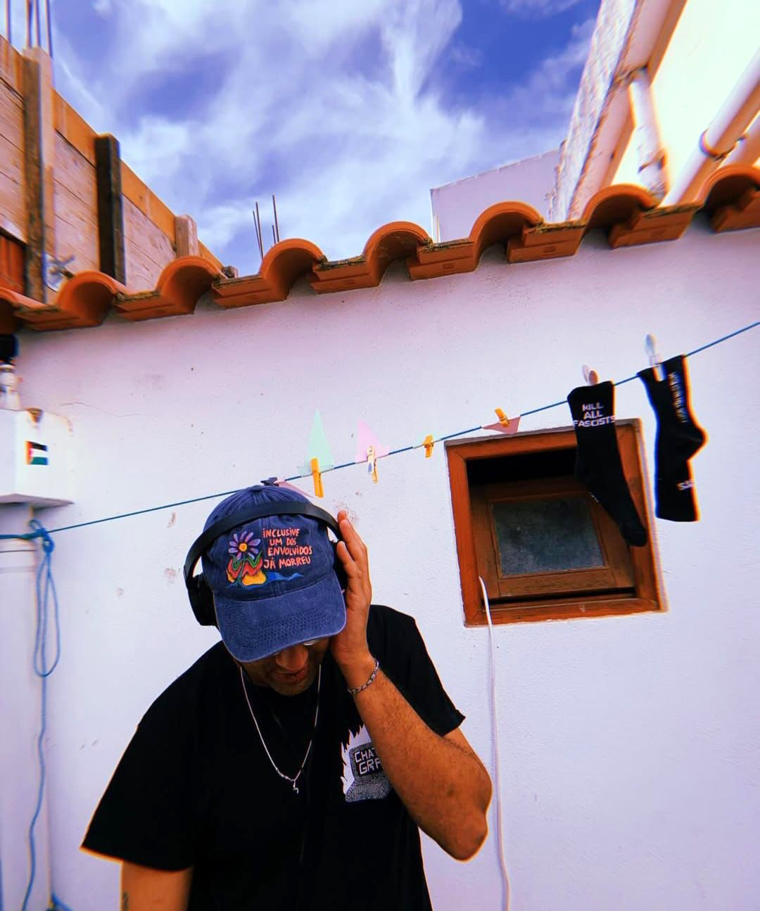 DJ Kali com auscultadores e boné, em frente a uma parede branca com roupa estendida