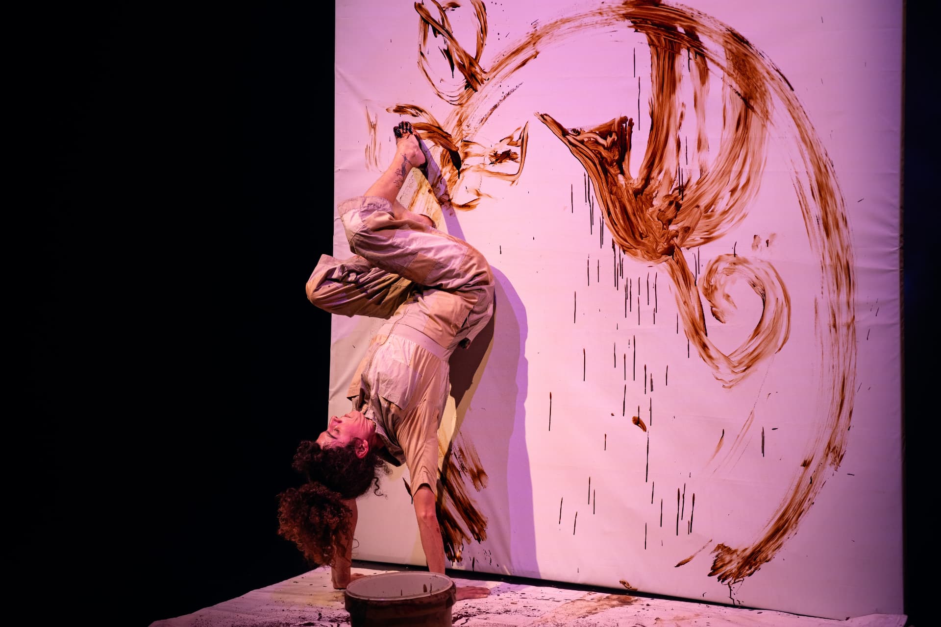 Lucia Merlina em equilíbrio invertido enquanto pinta com café sobre uma grande tela rosa